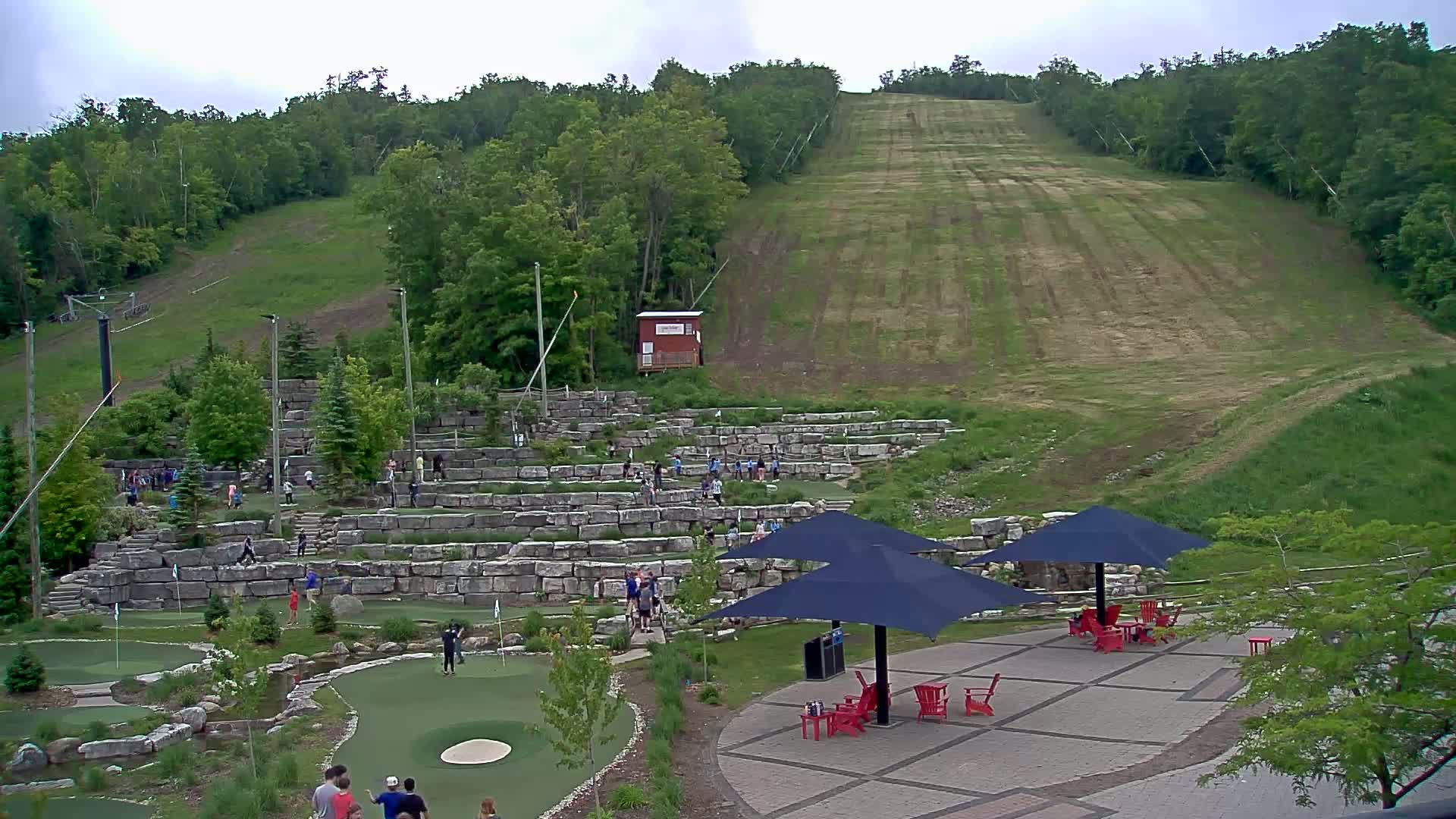 An outdoor mini golf course with people playing, situated below a grassy hill and surrounded by trees under an overcast sky.