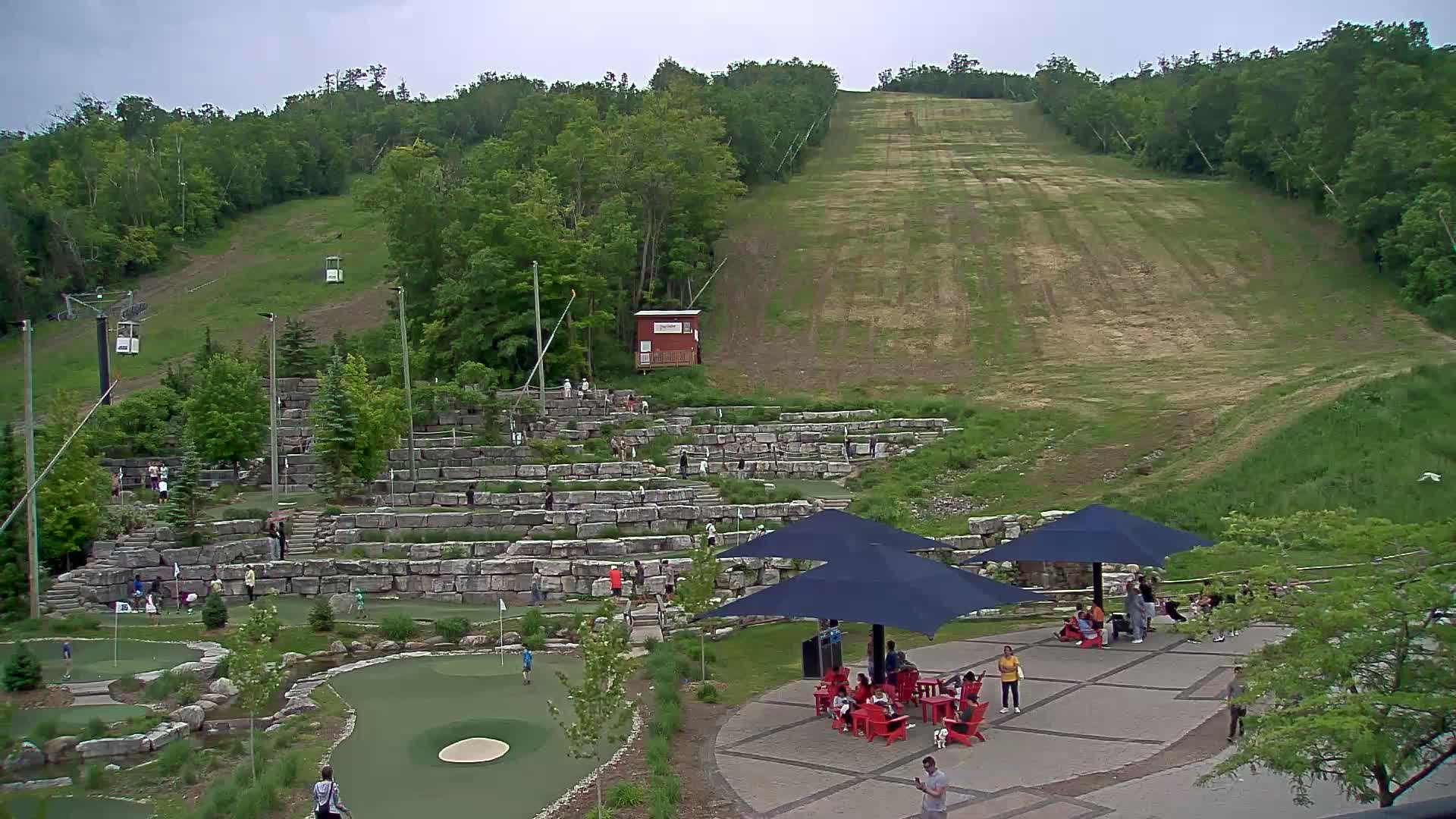 A grassy hill with a ski lift and a large stone structure with people playing mini golf, underneath an overcast sky.
