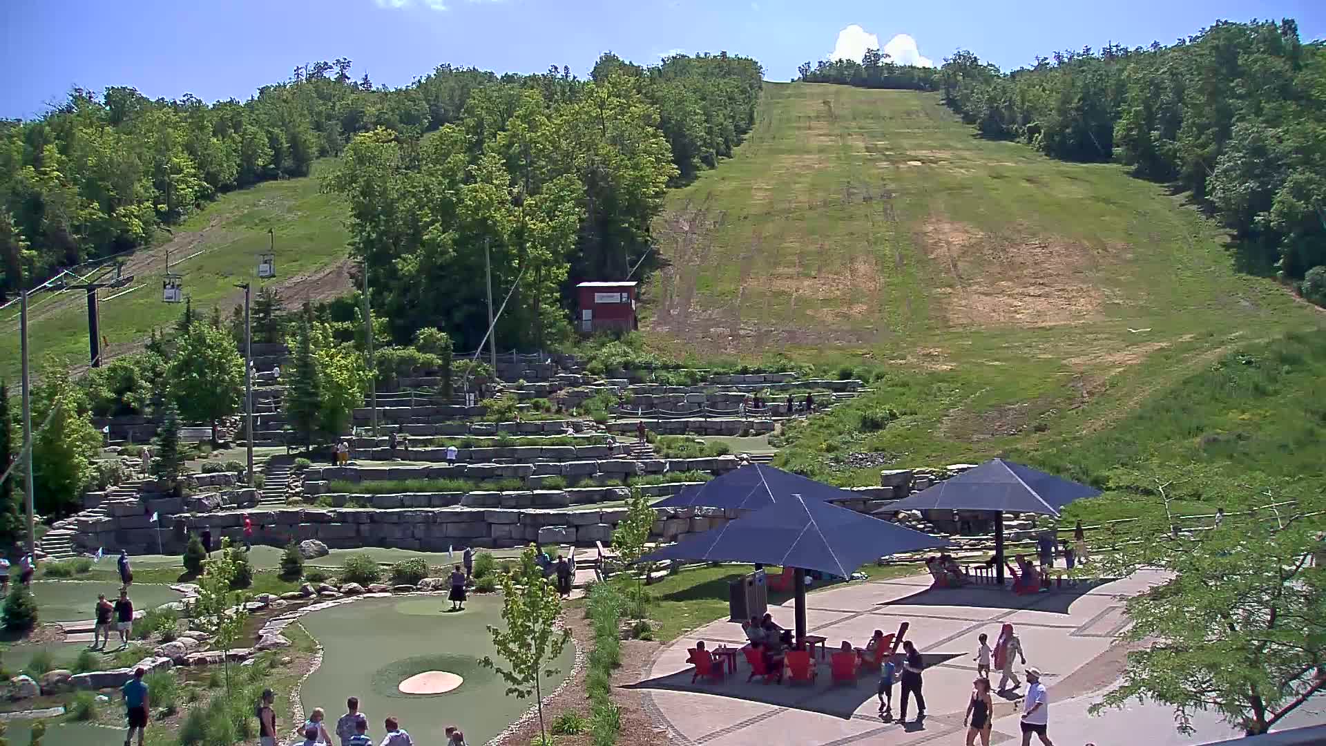 A sunny day shows a hillside with a ski lift and grassy slope, overlooking a paved area with people relaxing under umbrellas near a stone mini-golf course.