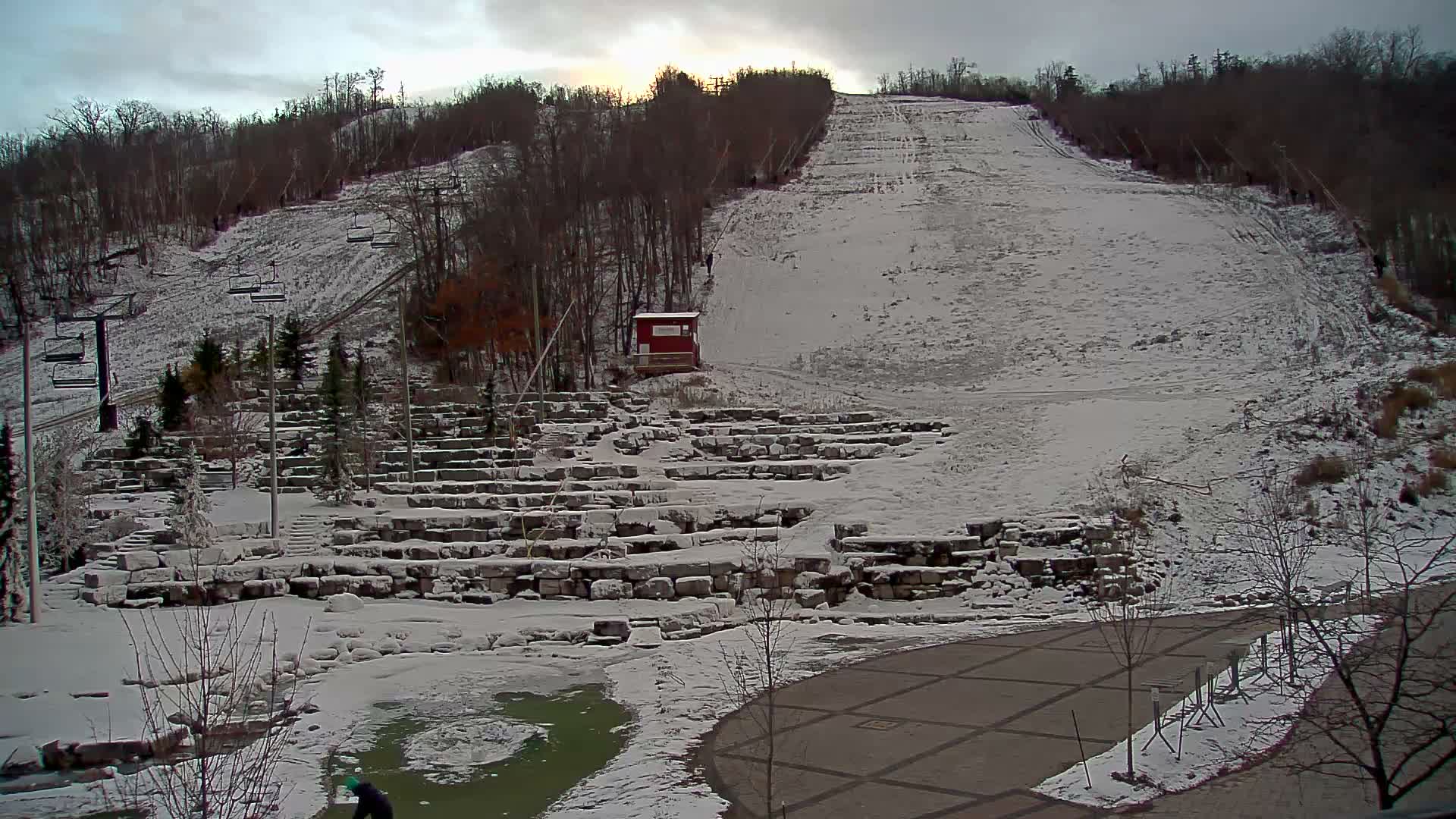 A snow-dusted ski slope and terraced rock garden are visible under an overcast sky with a faint sun peeking through, while a partially frozen pond and paved path fill the foreground, indicating cold, cloudy weather.