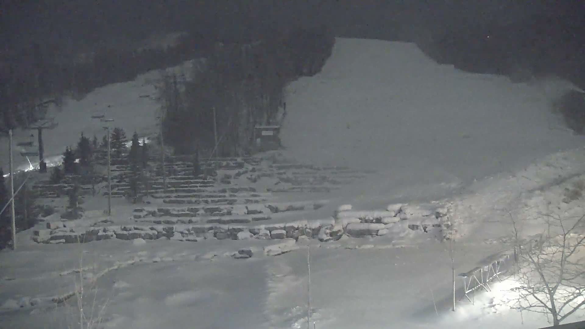 A snow-covered ski resort is seen at night under hazy conditions with light snow falling, featuring a prominent ski lift, numerous snow-dusted trees, and tiered rock formations.