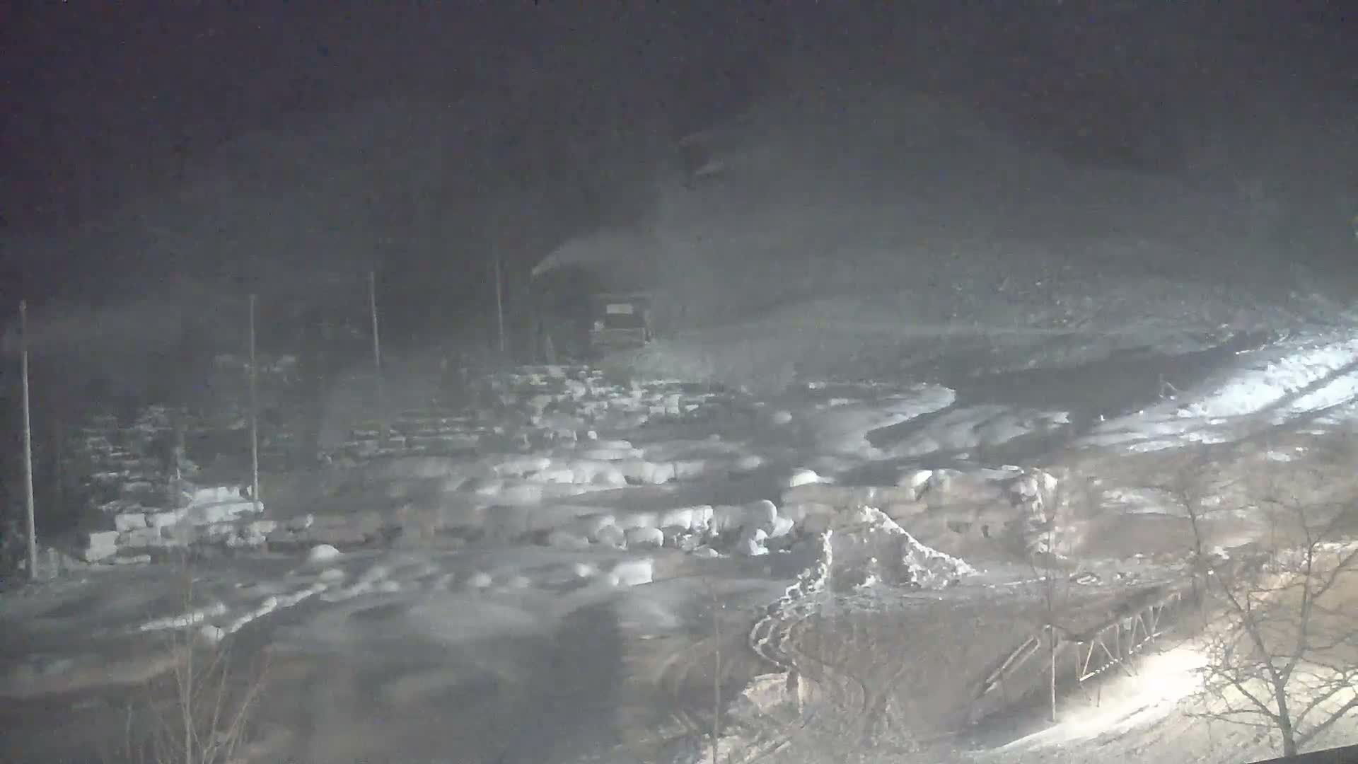 A hazy and cold night scene depicts a snow-making machine actively spraying snow over a snow-covered terrain featuring numerous mounds, bare trees, and an illuminated ramp-like structure in the foreground.