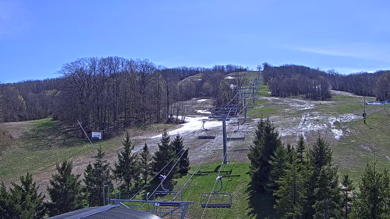 A chairlift ascends a grassy hill dotted with patches of melting snow under a clear blue sky.