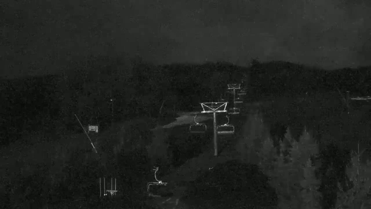 A nighttime view of a chairlift ascending a dark, wooded hill under a cloudy sky.