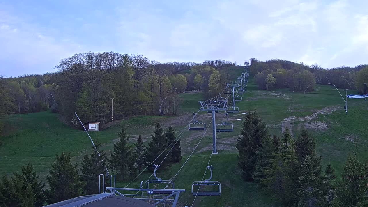 A chairlift ascends a grassy hillside covered in deciduous trees under a mostly clear, light blue sky.