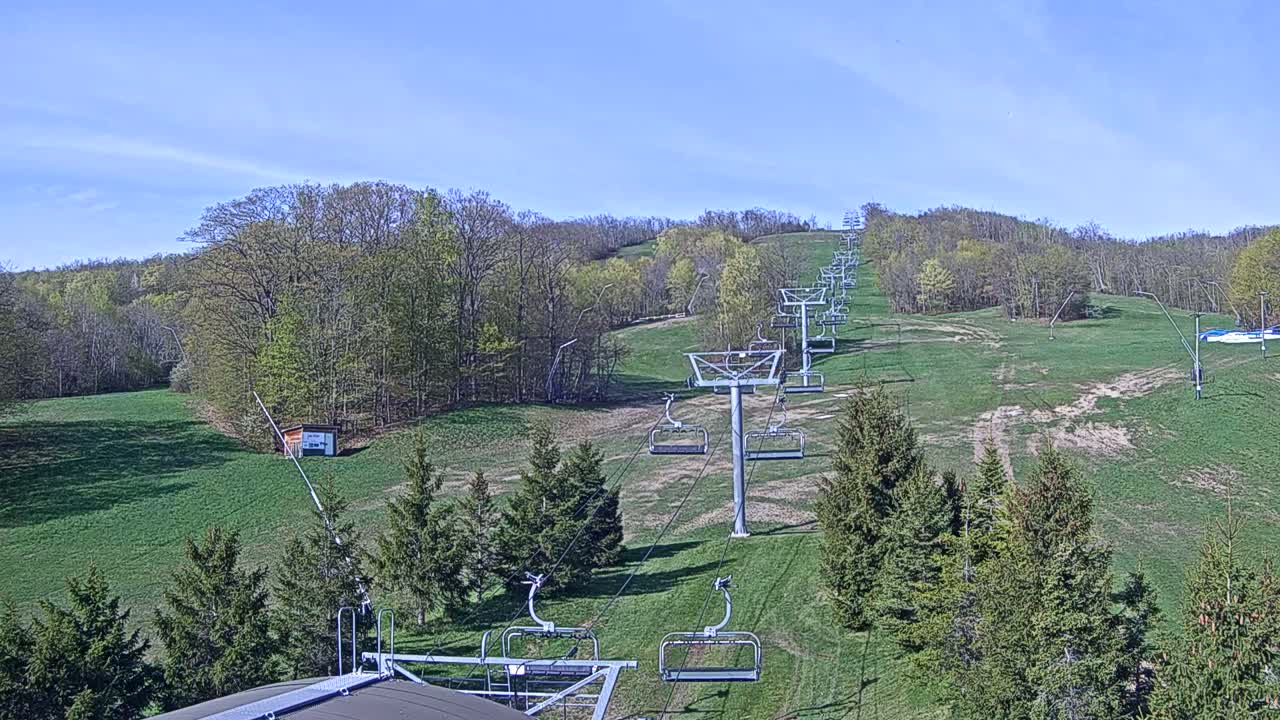 A chairlift ascends a grassy hill surrounded by budding deciduous trees under a clear blue sky.