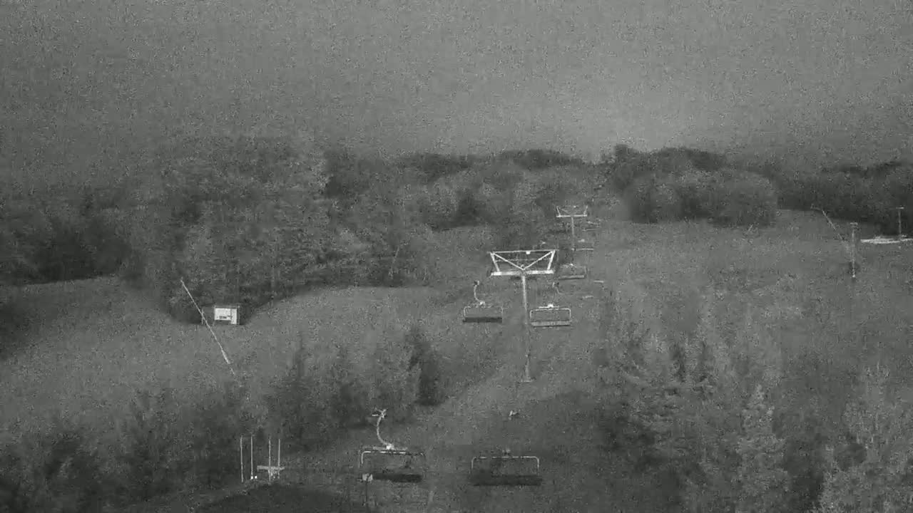 A ski lift with empty chairs is seen on a hill covered in trees under an overcast sky.