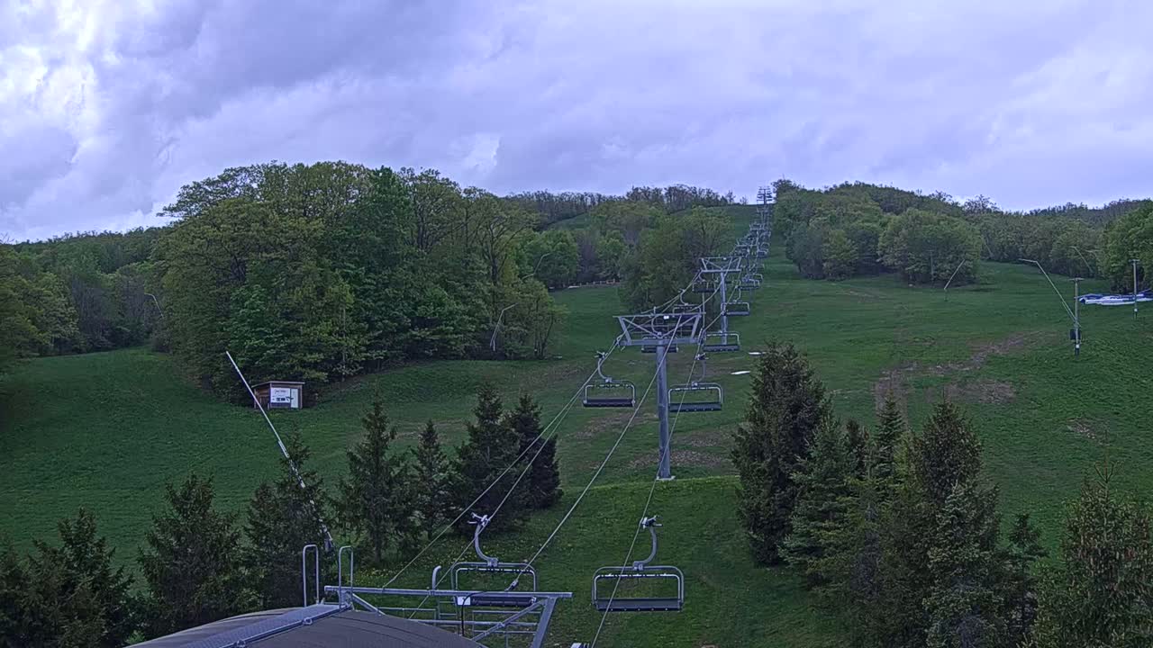 A chairlift ascends a grassy hill covered in green deciduous trees under a partly cloudy sky.