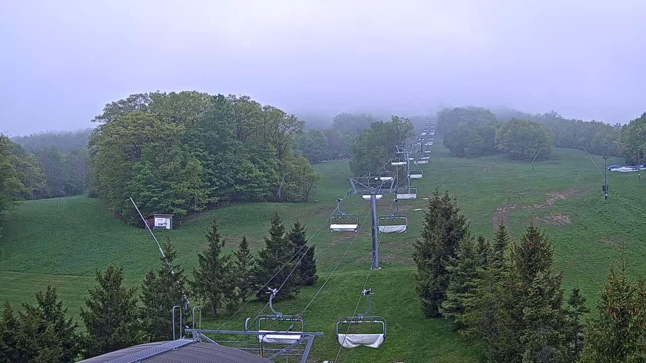 A chairlift stretches across a grassy hill shrouded in fog, with green trees lining the hillsides.