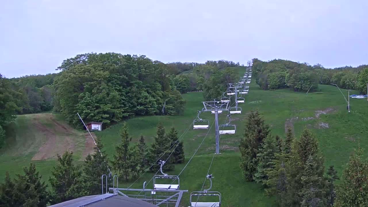 An empty chairlift ascends a grassy hill covered in green deciduous trees under an overcast sky.