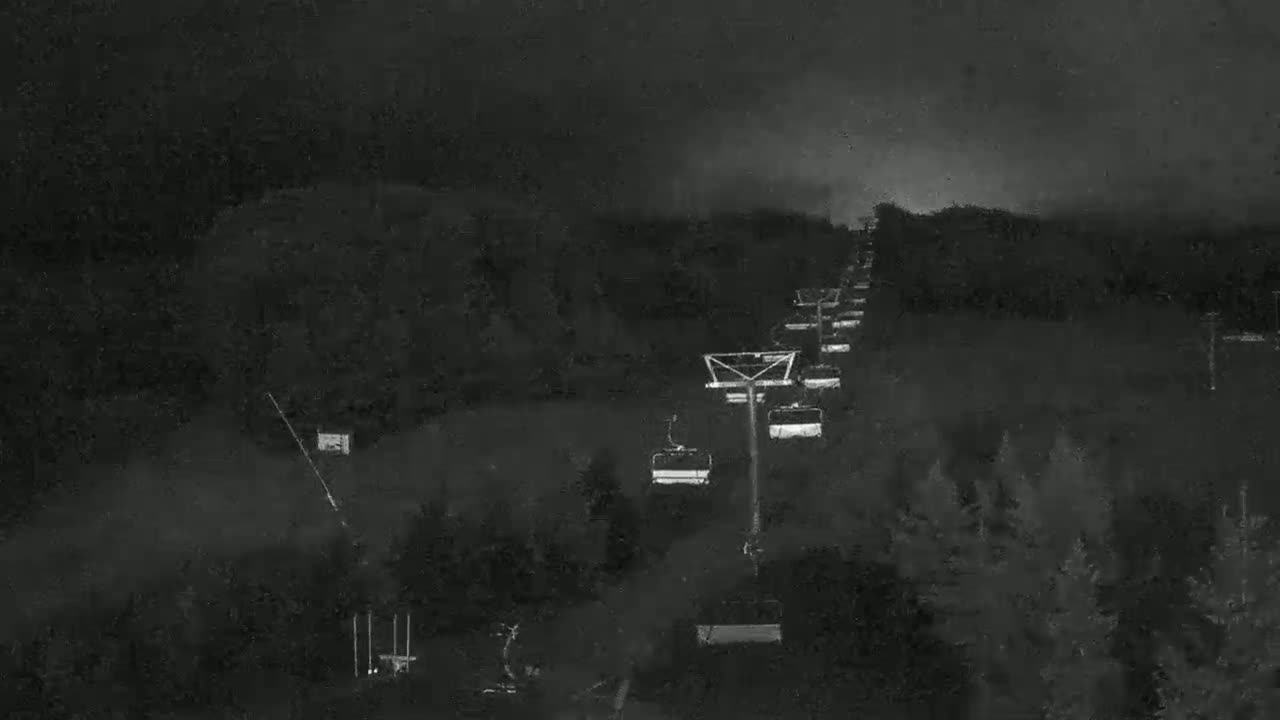 A nighttime view of a chairlift ascending a dark, wooded hillside under a cloudy sky.