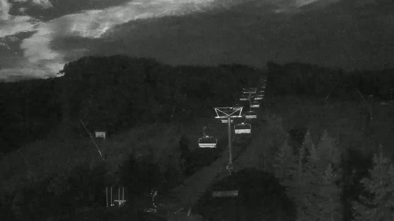 A chairlift ascends a dark, wooded hillside under a cloudy, nighttime sky.