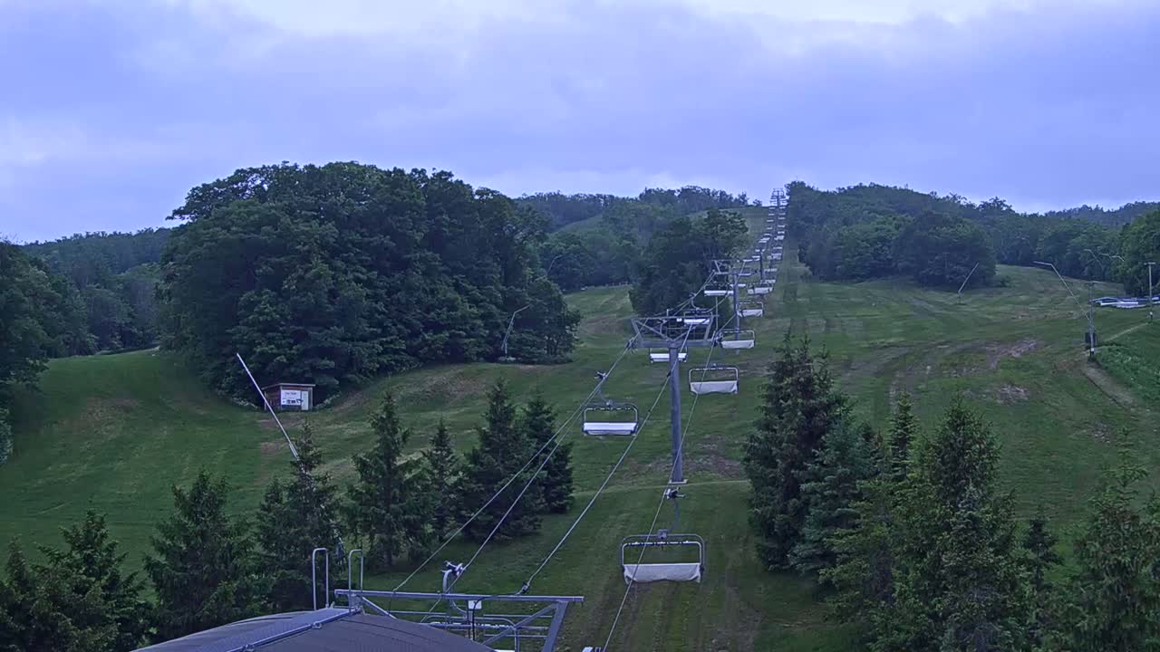 An empty chairlift ascends a grassy hill covered in green trees under a cloudy sky.