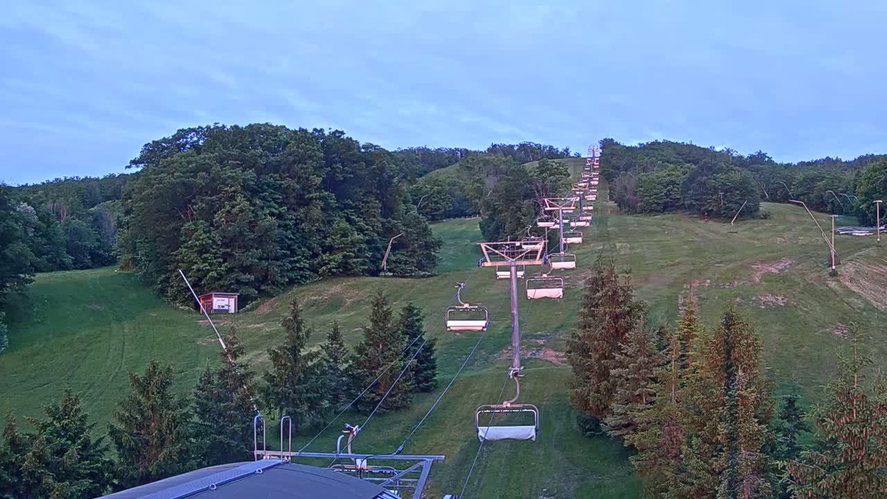 An empty chairlift ascends a grassy hill dotted with evergreen trees under a clear twilight sky.