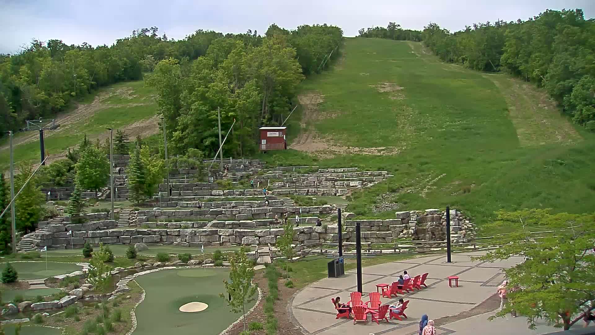 A grassy hillside with a ski lift rises behind a stone terraced area containing a mini golf course and people relaxing in red chairs on a patio under a partly cloudy sky.
