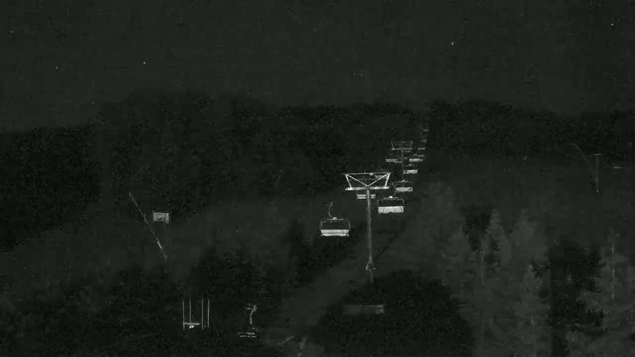 A nighttime view of a chairlift ascending a dark, wooded hillside under a mostly clear sky.