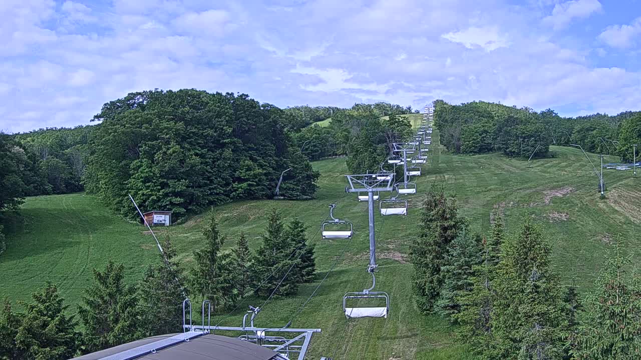 A chairlift ascends a grassy hill covered in lush green trees under a partly cloudy sky.