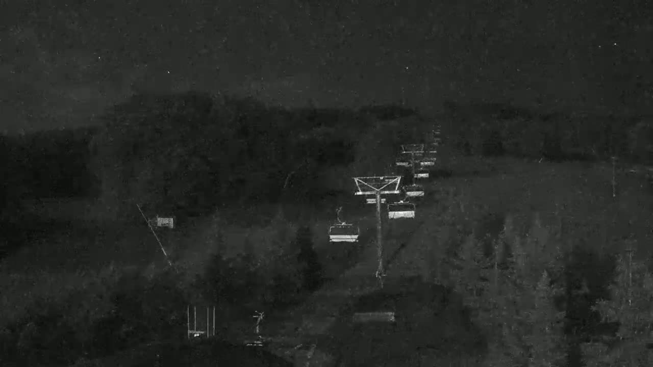 A nighttime view of a chairlift ascending a snow-covered hillside under a dark, possibly starry sky.