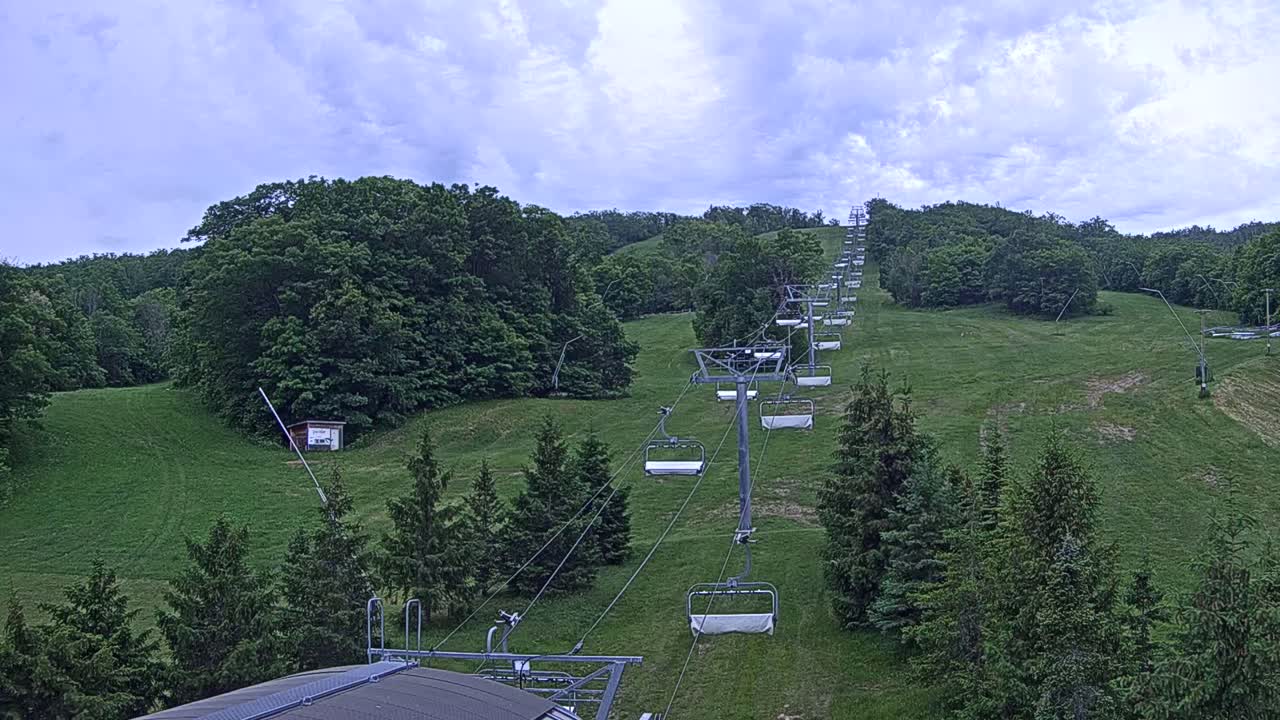 A chairlift ascends a grassy hillside covered in lush green trees under a partly cloudy sky.