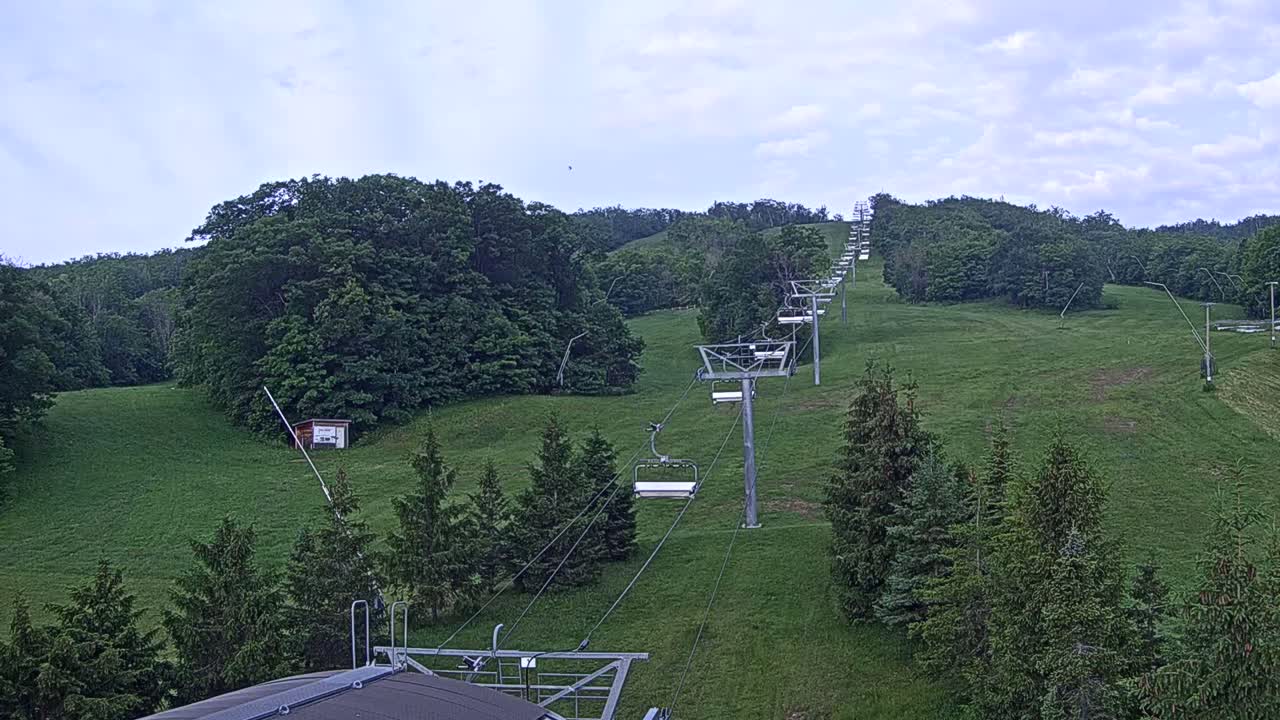 A chairlift ascends a grassy hill covered in green trees under a partly cloudy sky.