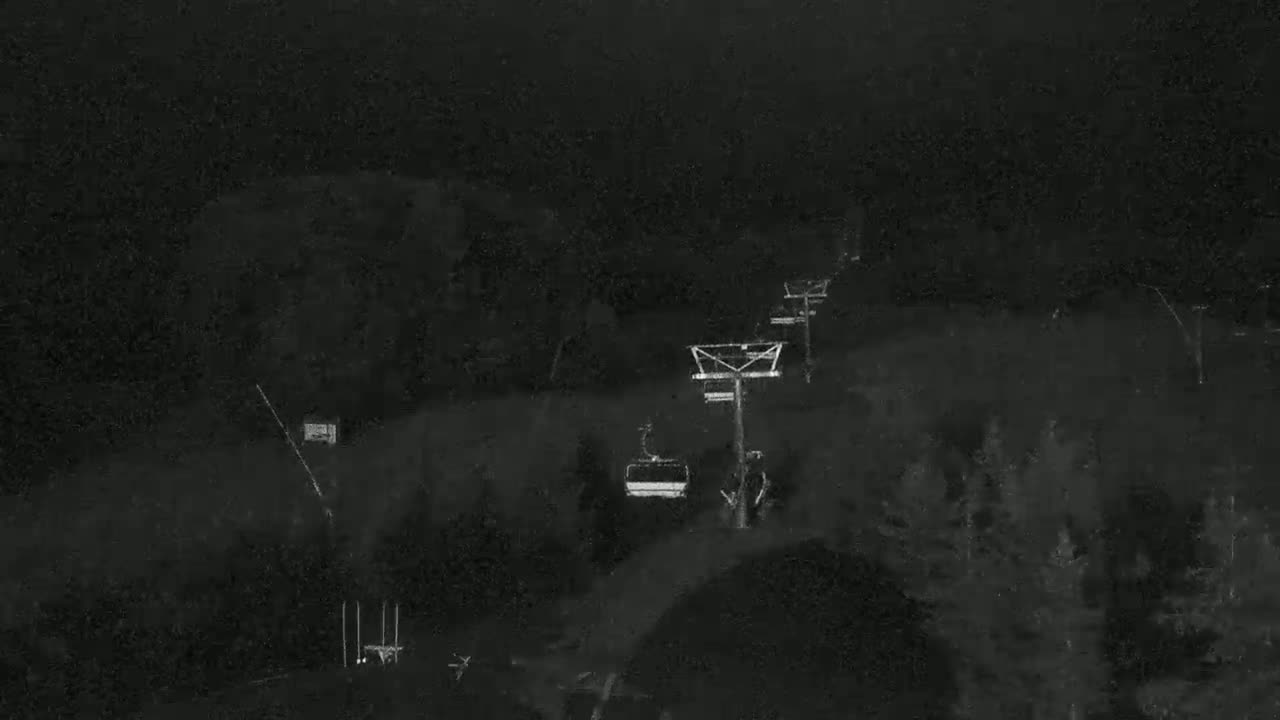 A nighttime view of a ski lift with a single chairlift moving across a snow-covered hillside under a dark, possibly snowy sky.