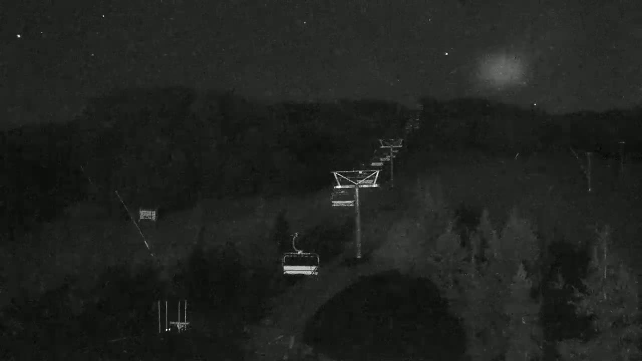 A chairlift ascends a dark, wooded hillside under a cloudy, nighttime sky.
