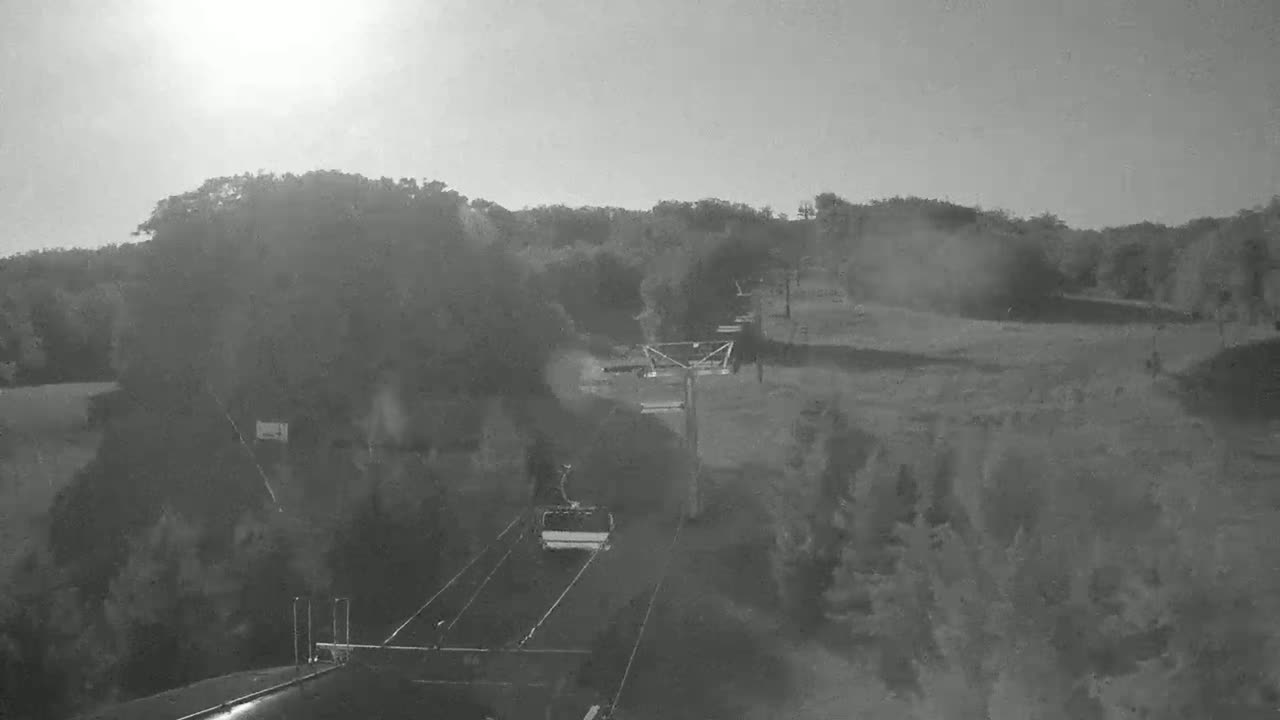 A chairlift carrying a single passenger moves along a track through a hazy, sunlit landscape of trees and open fields.