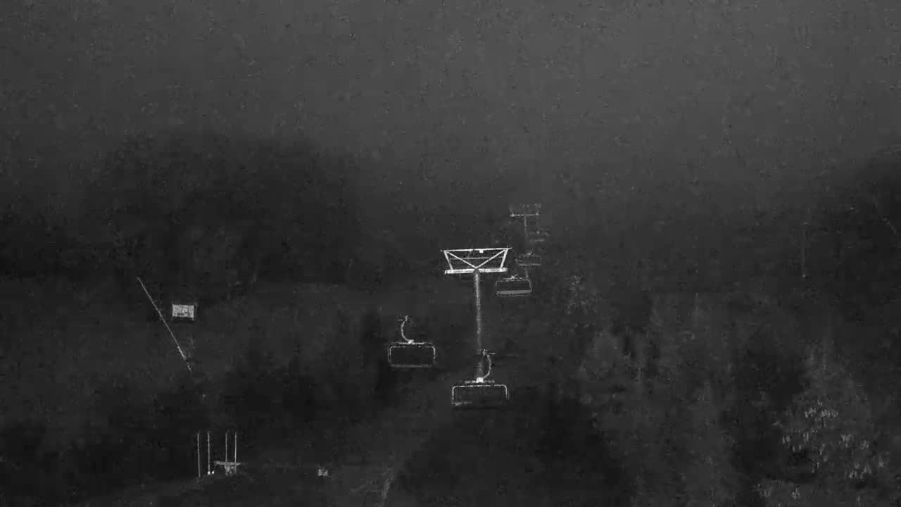 A dark, grainy, monochrome view of a ski slope features a ski lift tower and several empty chairs ascending through what appears to be heavy fog or night with widespread precipitation.
