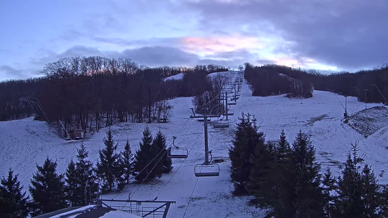 A snow-covered ski slope with empty chairlifts ascending a forested mountain is visible under an overcast sky showing subtle pink and blue hues, characteristic of dawn or dusk.