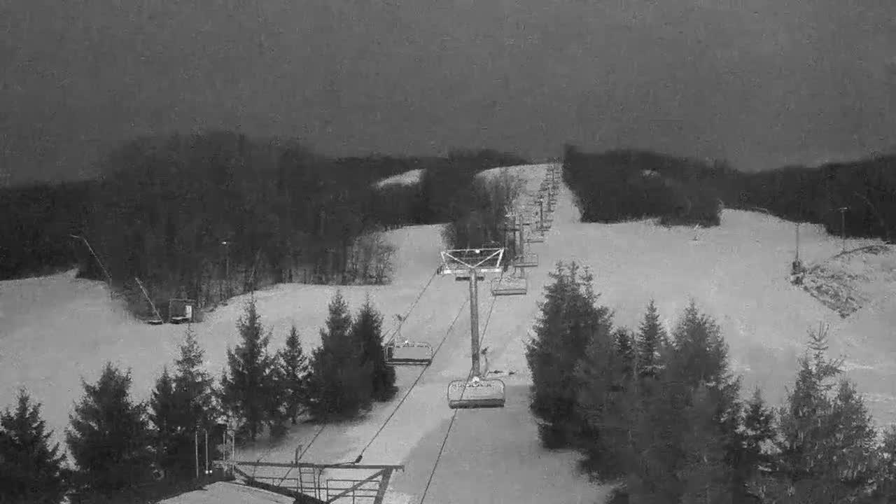 A snow-covered ski slope featuring an empty chairlift ascending uphill, bordered by evergreen trees, is visible under a uniformly overcast winter sky.