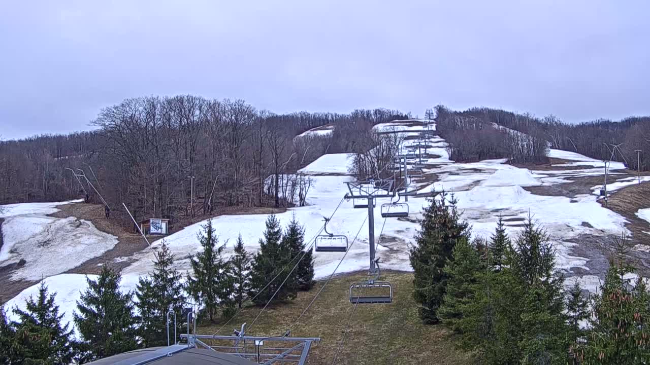 A snow-covered ski slope featuring an empty chairlift ascending uphill, bordered by evergreen trees, is visible under a uniformly overcast winter sky.
