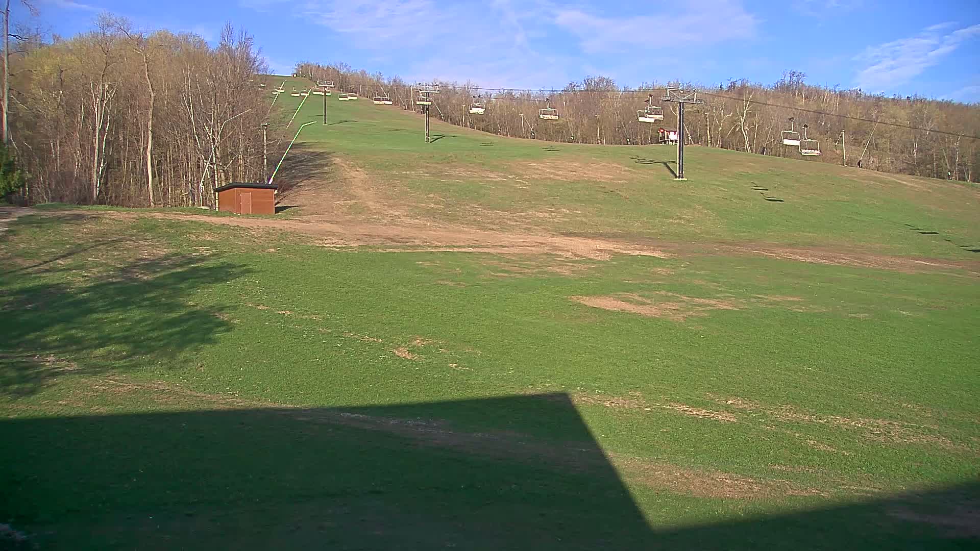 A grassy ski slope with empty chairlifts under a mostly sunny sky.