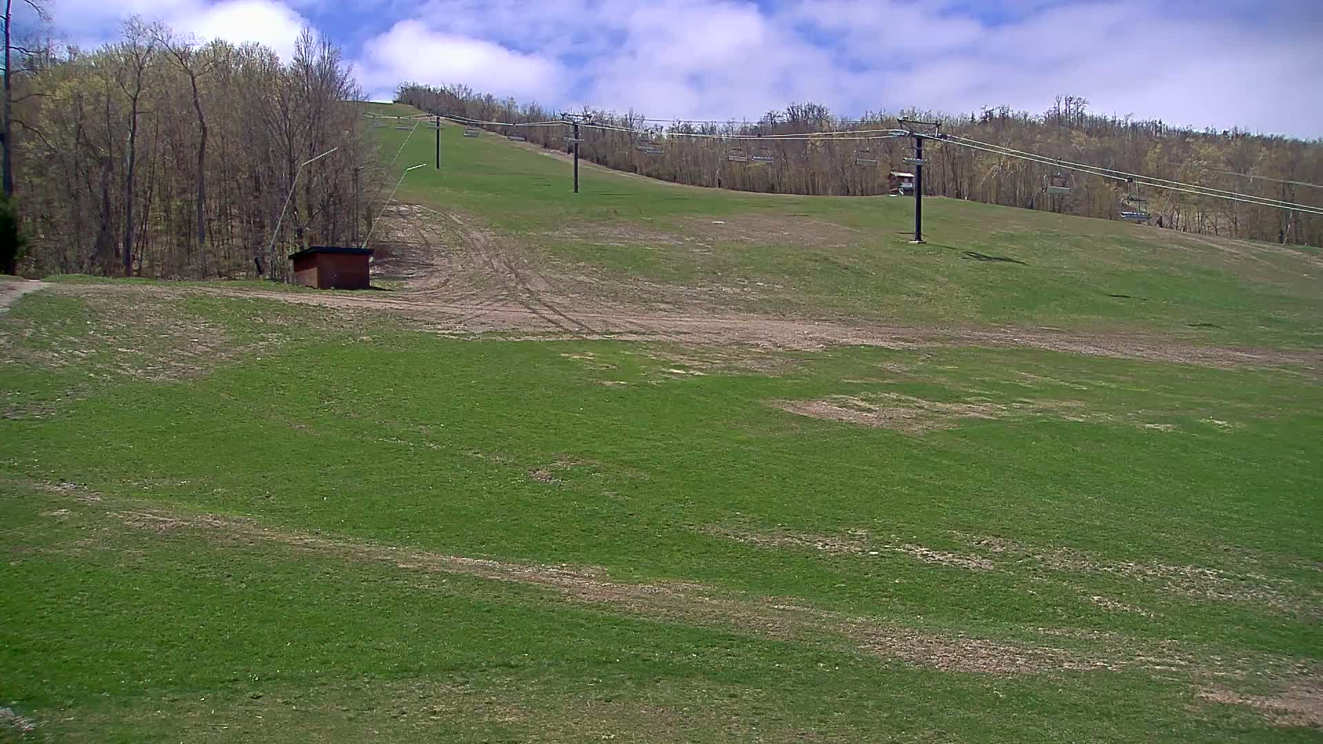 A grassy hillside with a ski lift and a small building under partly cloudy skies.