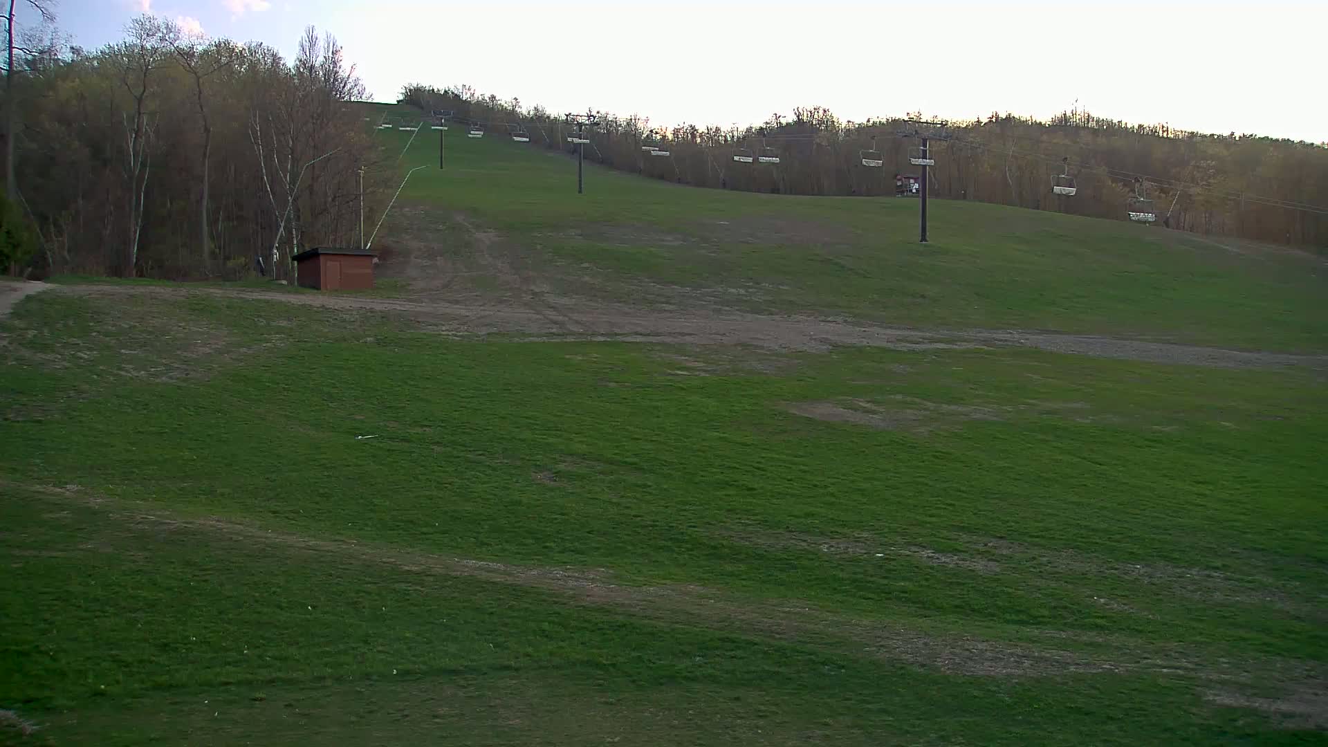 A grassy ski slope with empty chairlifts and a small building at the bottom, under a partly cloudy sky.