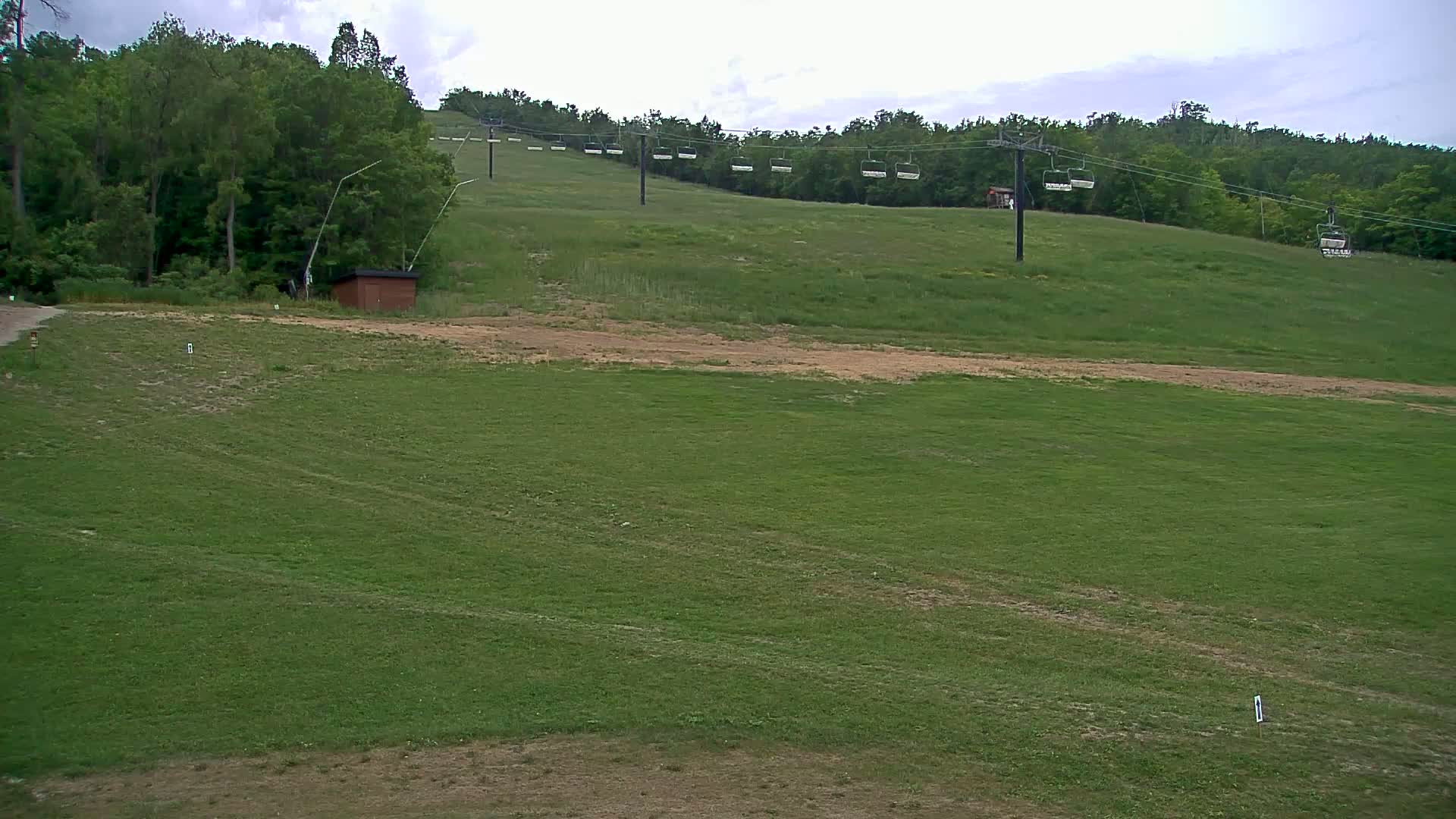 A grassy hillside with an empty chairlift and a small brown building under a partly cloudy sky.