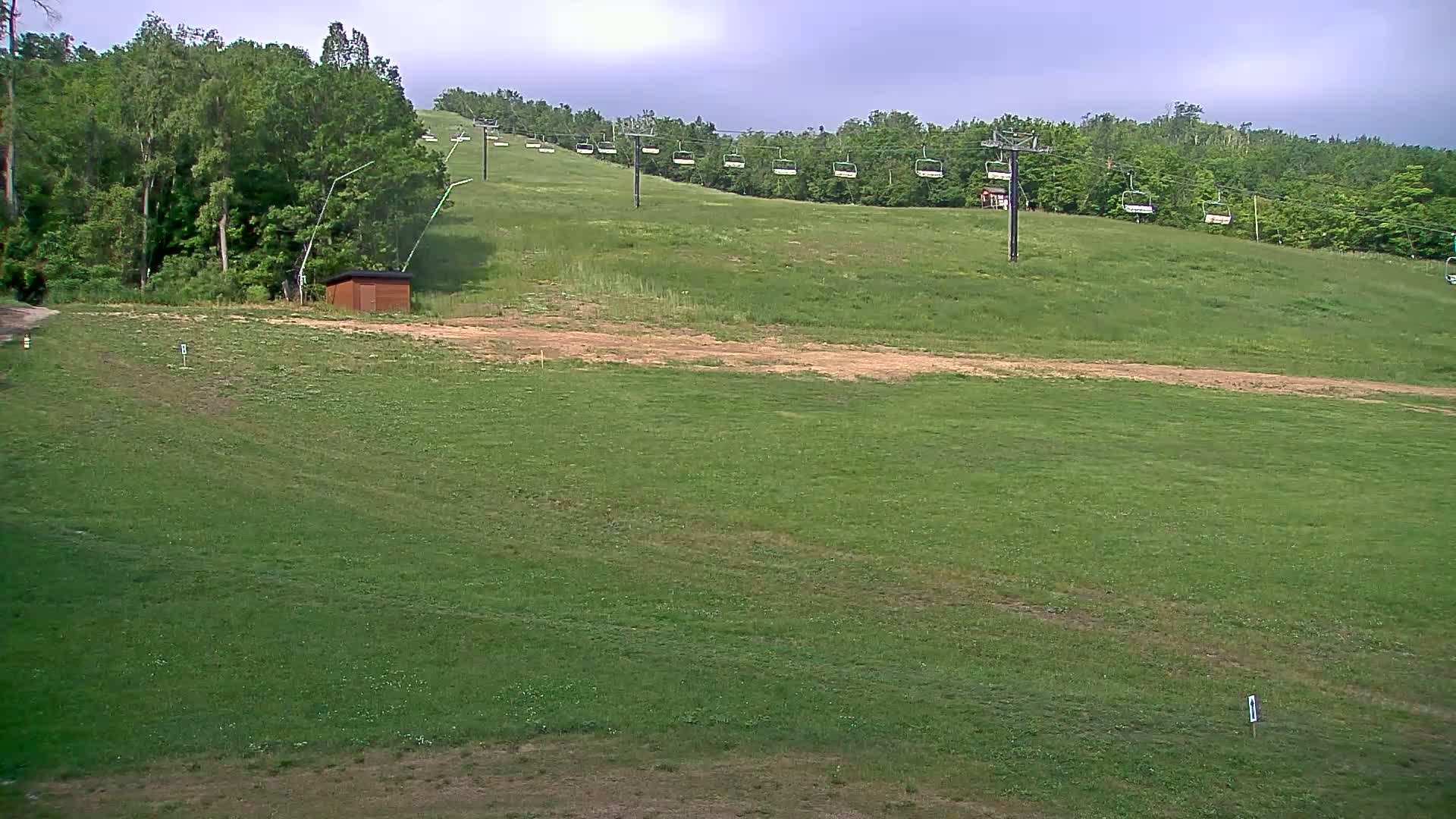 A grassy hill with a chairlift and a small shed under a mostly cloudy sky.