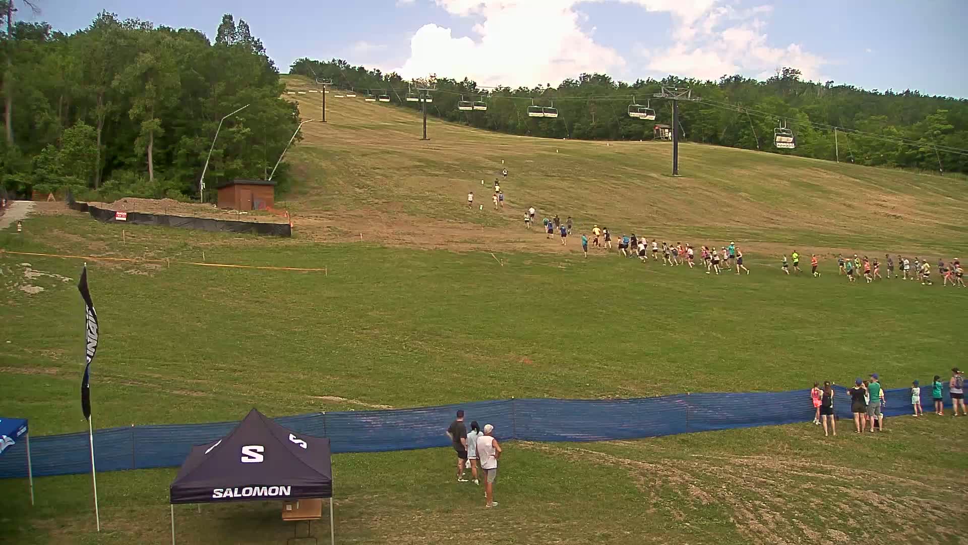 A large group of people runs up a grassy hill under a partly cloudy sky, with a smaller group of spectators near the bottom of the hill.