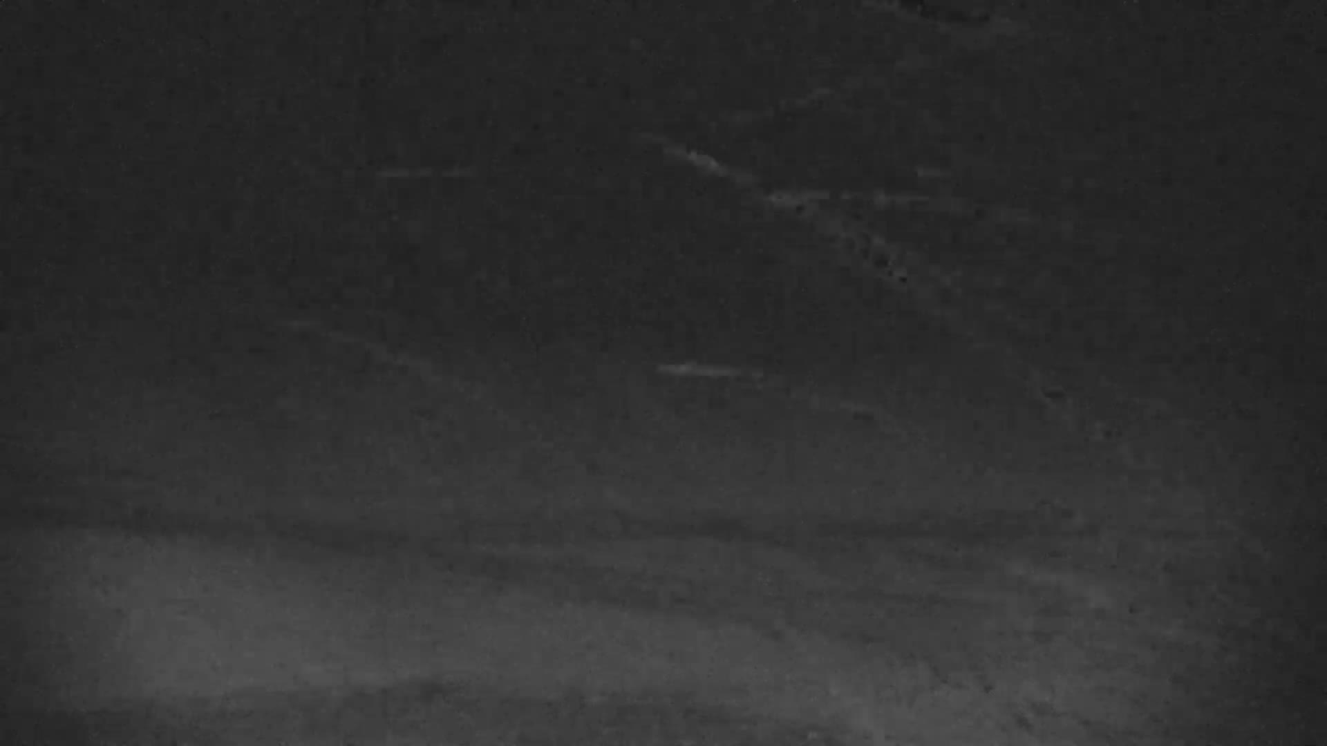 A dark, grainy, black and white image depicts an expansive, indistinct outdoor landscape at night, featuring subtle horizontal patterns suggesting a ground or water surface and faint, elongated bright forms under extremely low visibility conditions.