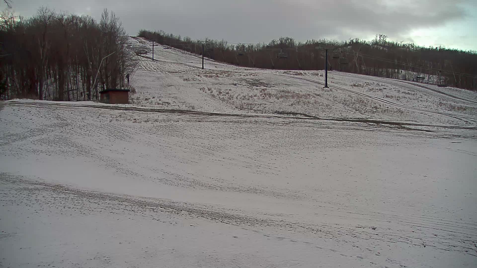 Snow-dusted ski slopes, featuring a small building, numerous bare trees, and empty ski lift towers and chairs, are visible under an overcast and cloudy sky.