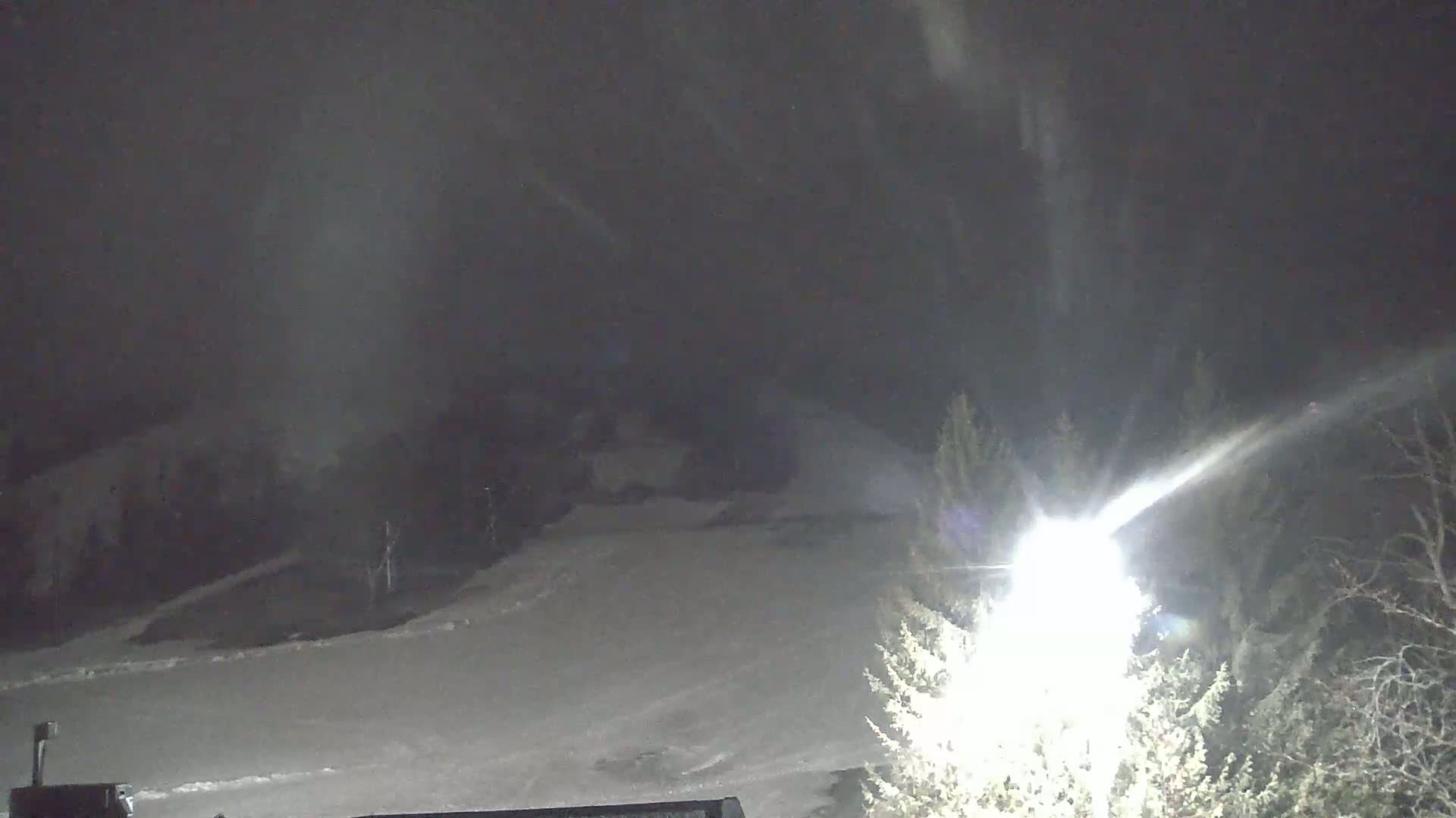A snow-covered landscape at night is illuminated by a bright light source, with snow falling in the distance.