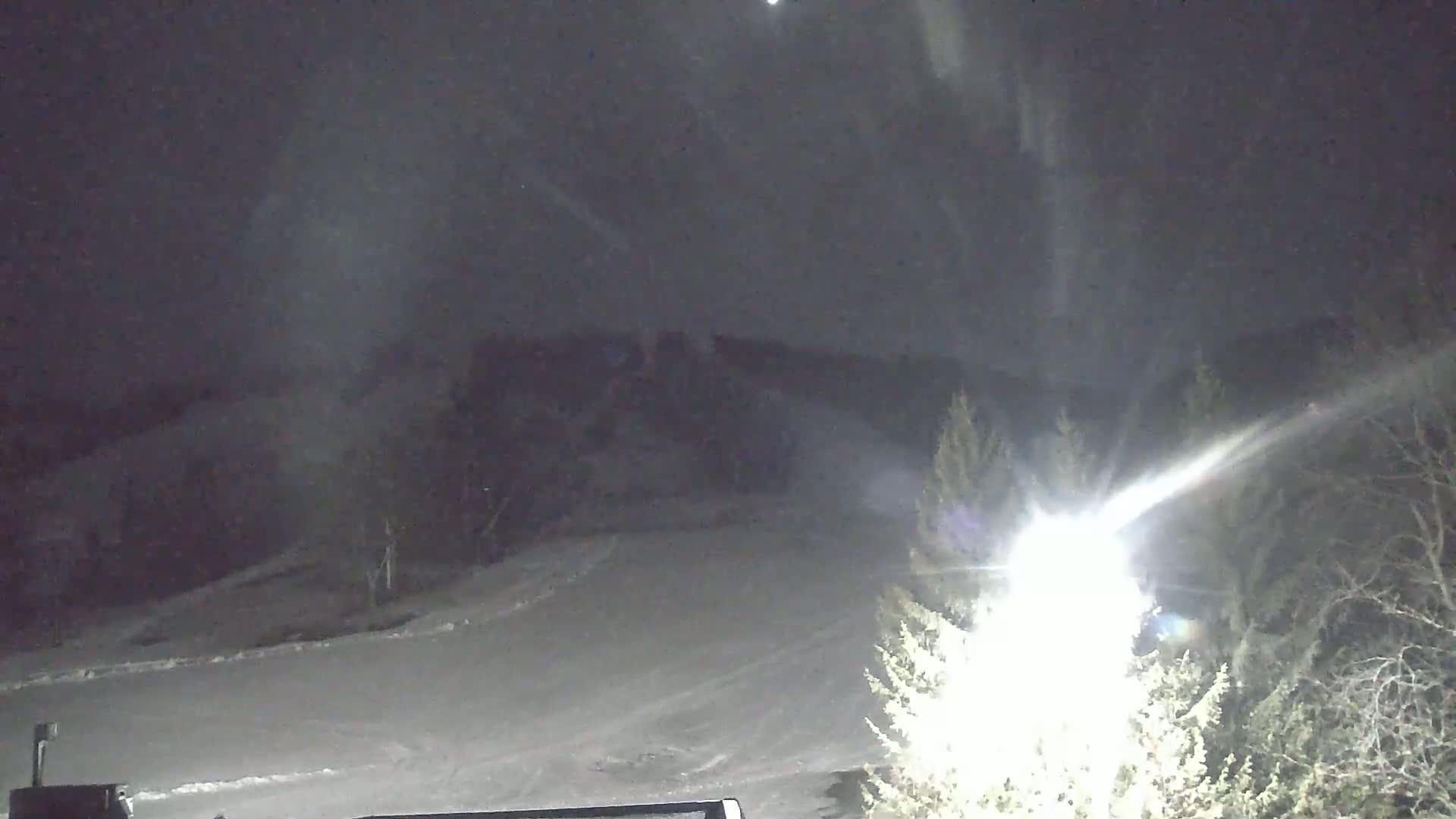 A snow-covered landscape is illuminated by a bright light at night, with snow falling lightly.