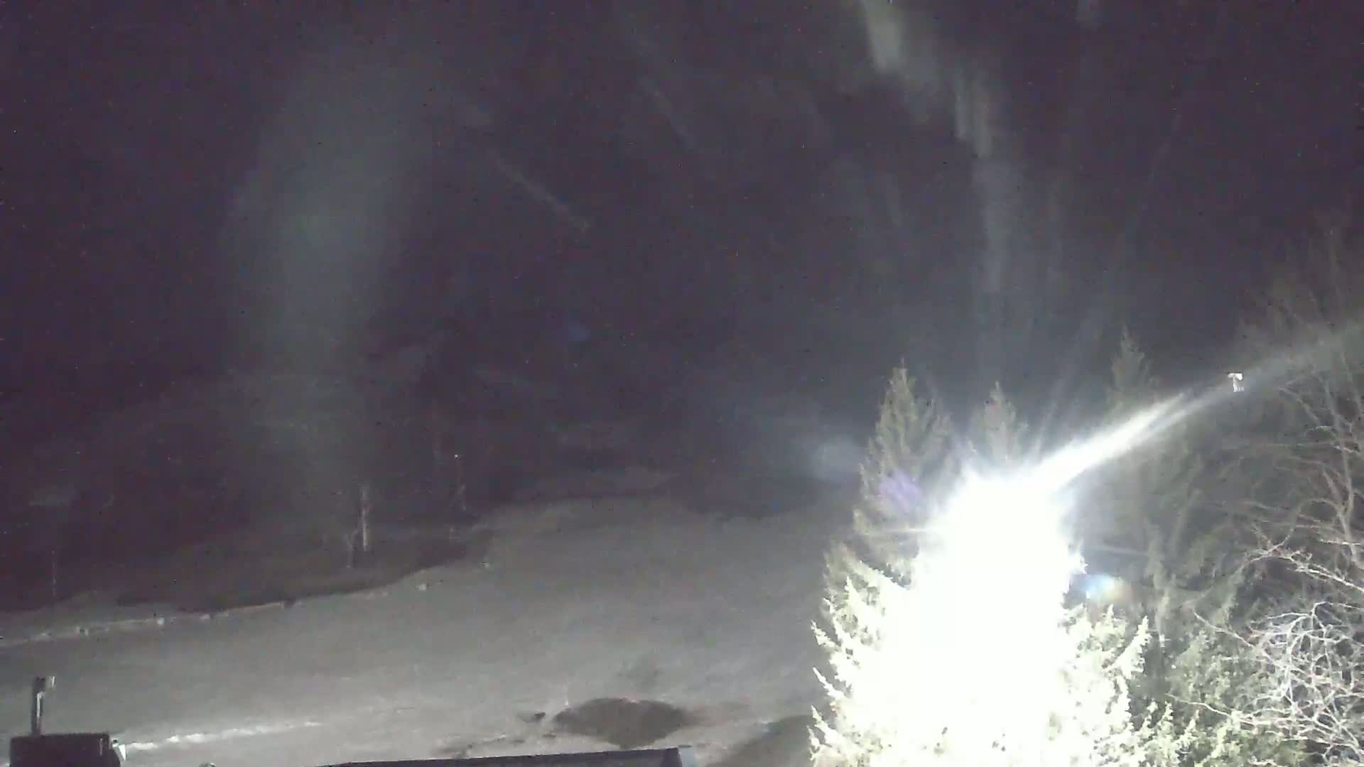 A snow-covered landscape at night is illuminated by a bright light source, with snow falling or blowing in the air.