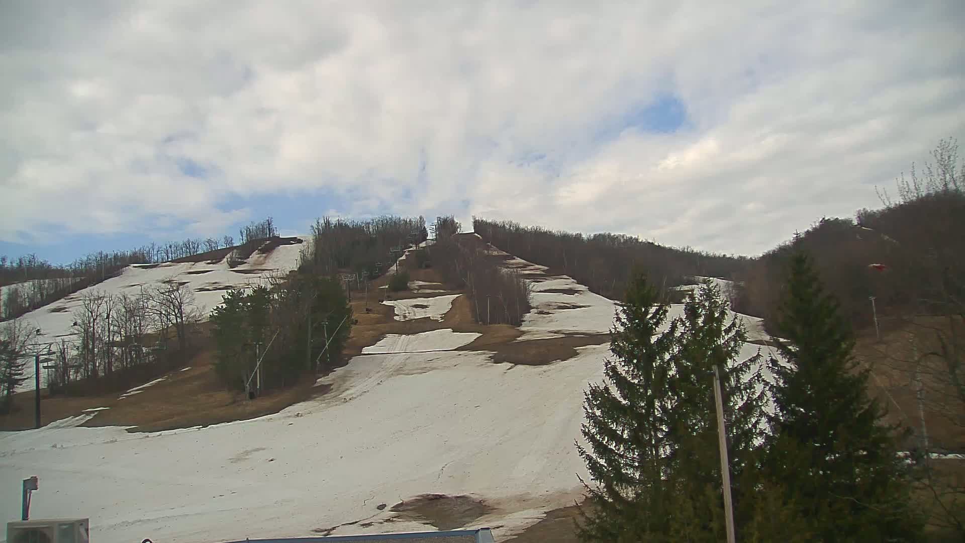 A mostly cloudy day reveals a ski hill with patches of snow and brown earth, interspersed with leafless trees and ski lifts.