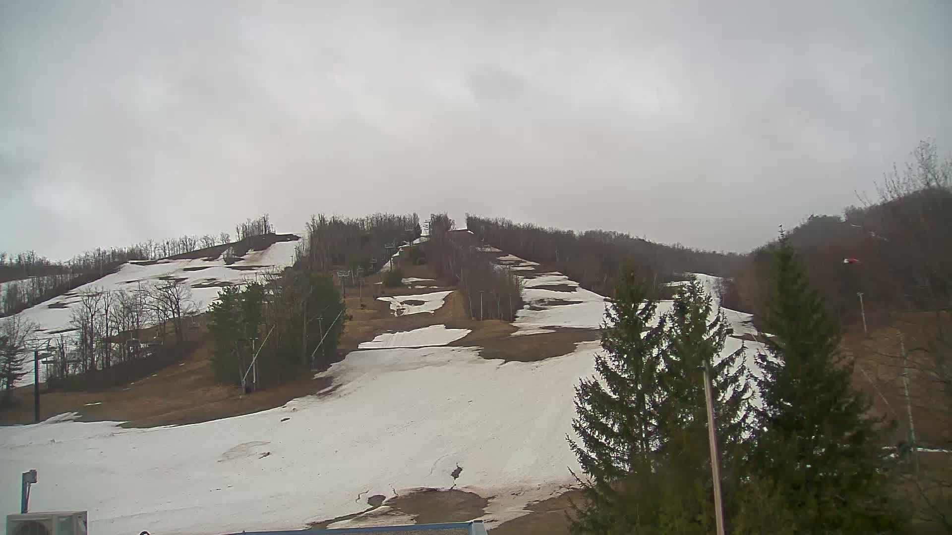 A mostly snow-covered ski hill with patches of brown ground, under an overcast sky.