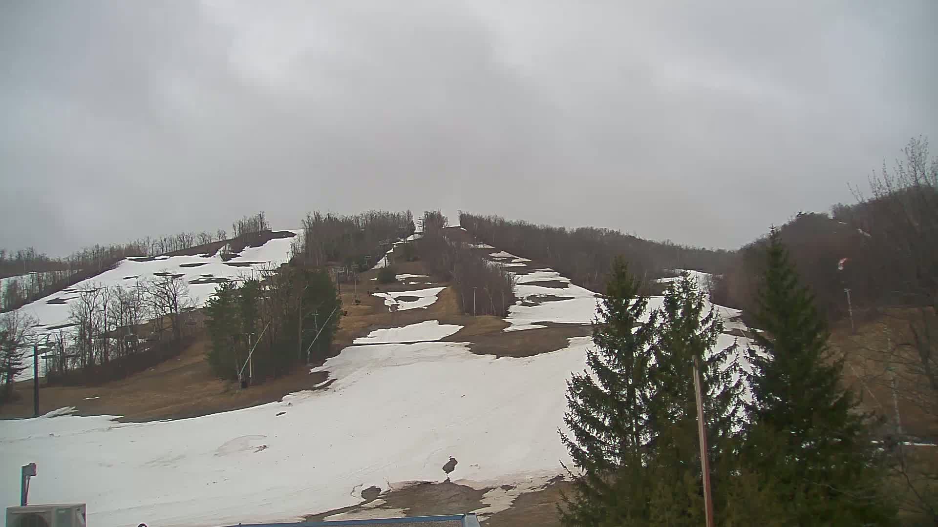 A snow-covered ski hill with patches of bare ground is visible under an overcast sky.