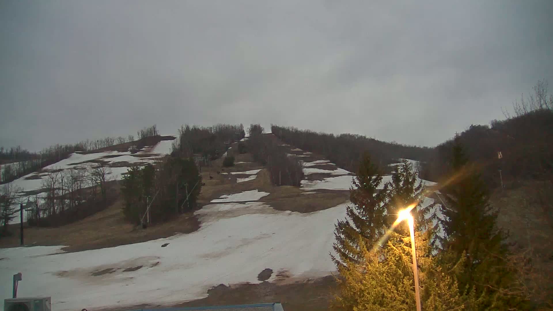 A snow-covered ski slope on a hill under an overcast sky, with evergreen trees in the foreground.