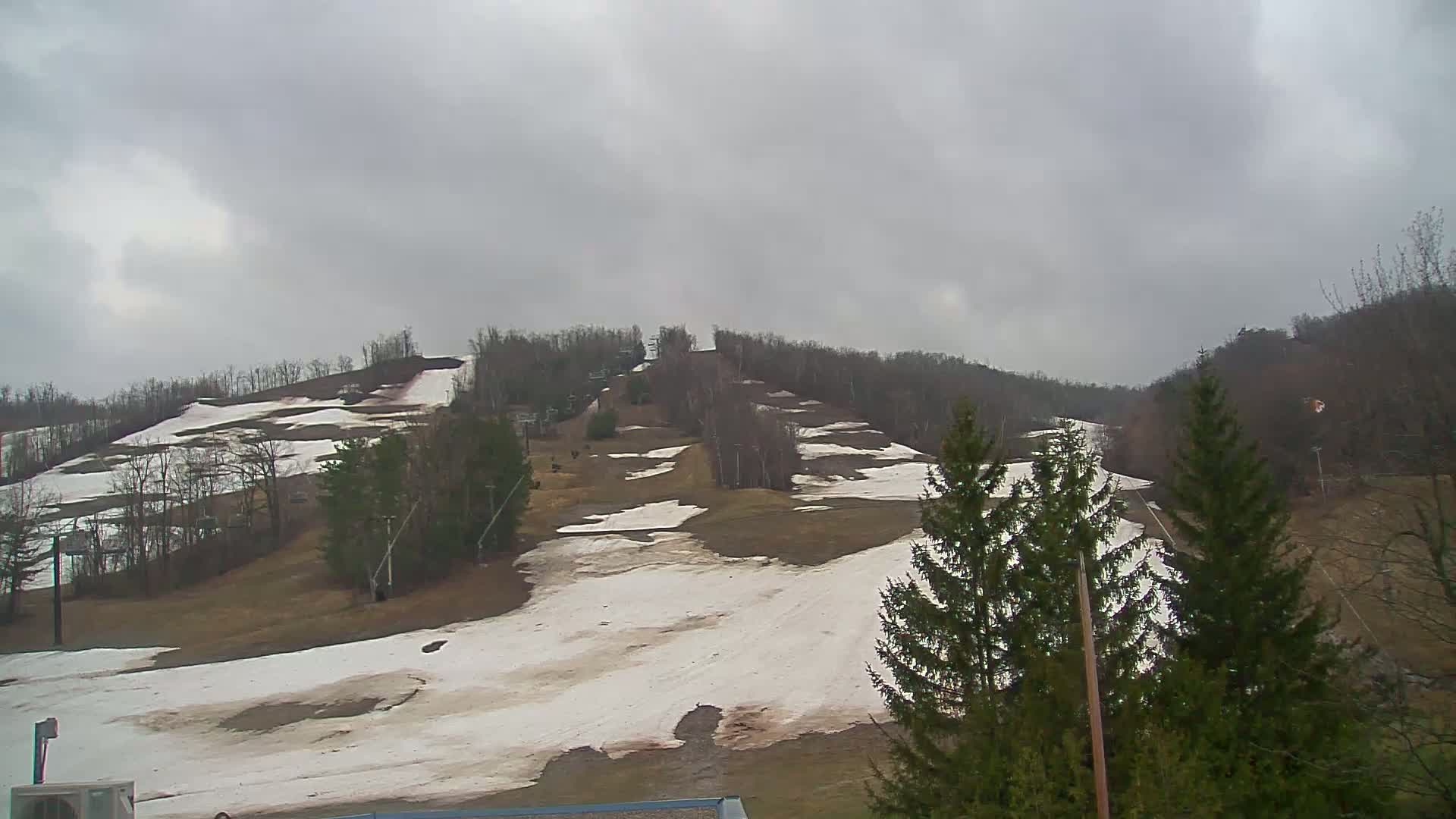 Patches of snow remain on a hillside ski slope under a cloudy sky.