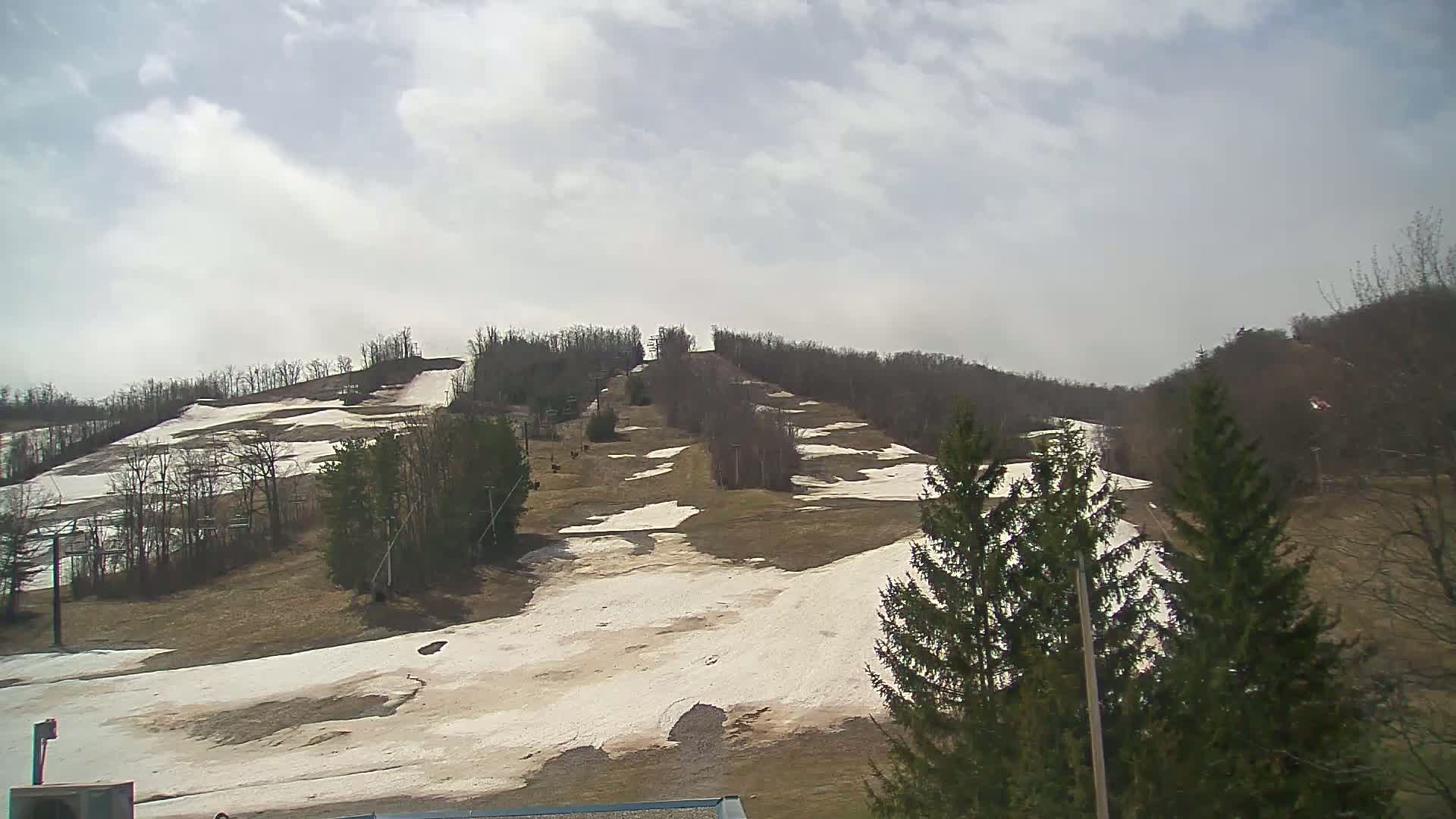A mostly cloudy day reveals a ski slope with patches of melting snow and brown grass on a hill covered in leafless trees.