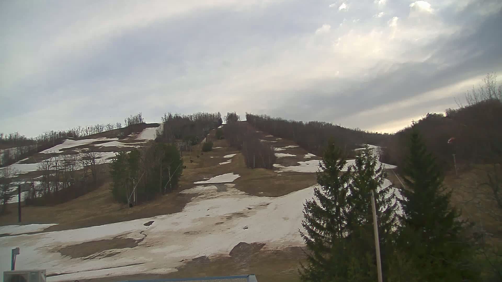 A mostly cloudy sky overlooks a ski hill with patches of melting snow and bare, brown grass interspersed among trees.