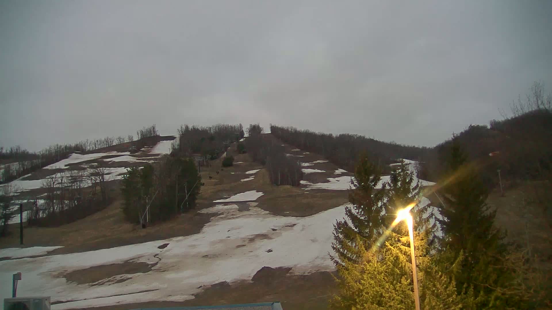 A mostly cloudy day reveals a ski hill with patches of snow and brown grass, illuminated by a streetlamp in the foreground.